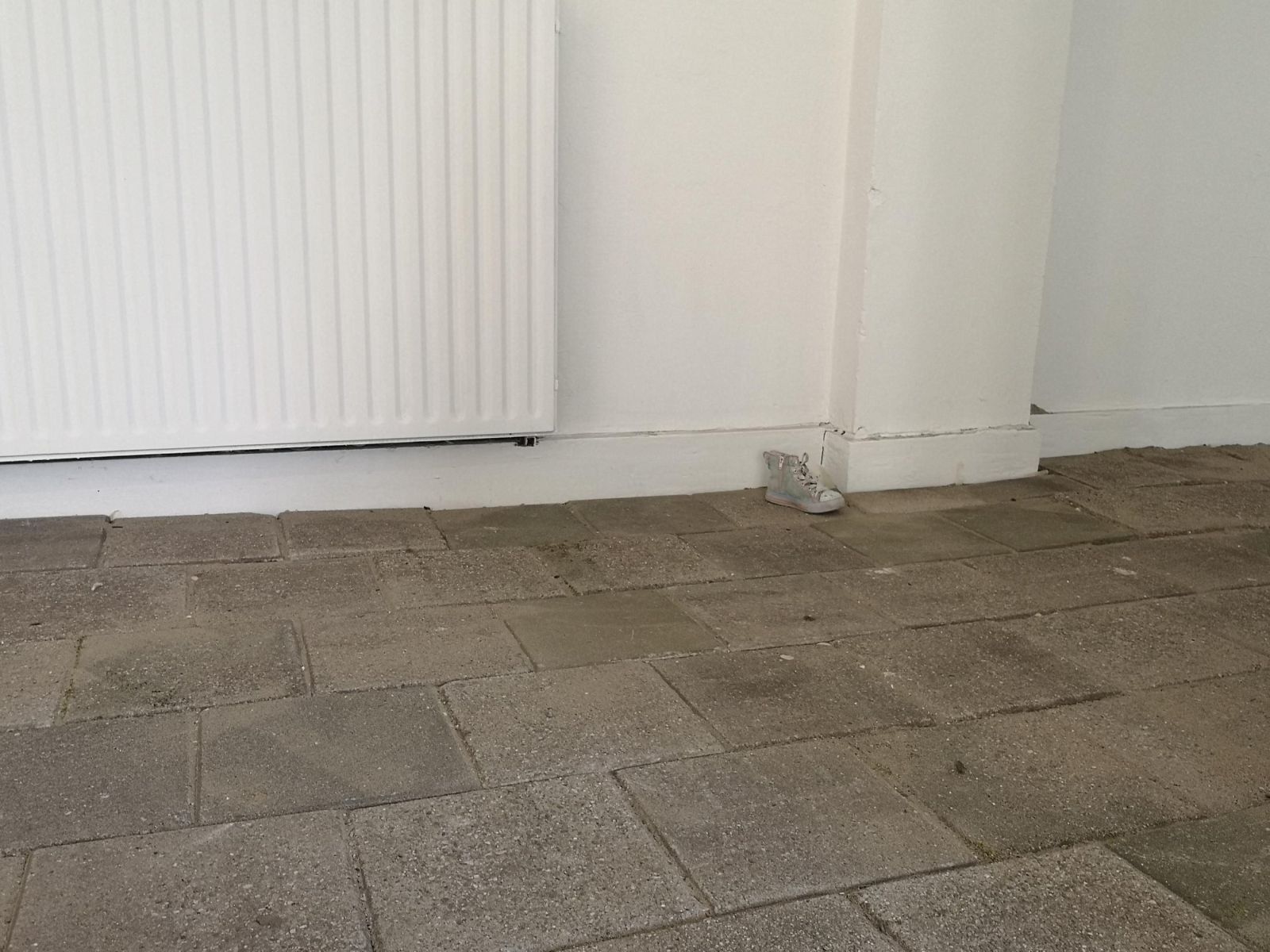 The ground is made of street tiles. In the background you see a white wall and radiator. Centre left a small pink and blue kids shoe lies on the ground