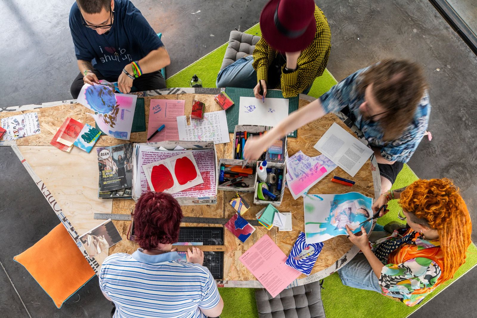 five people sitting around a weirdly shaped table. on the table are many different colorful sheets of paper and everyone is creating something.