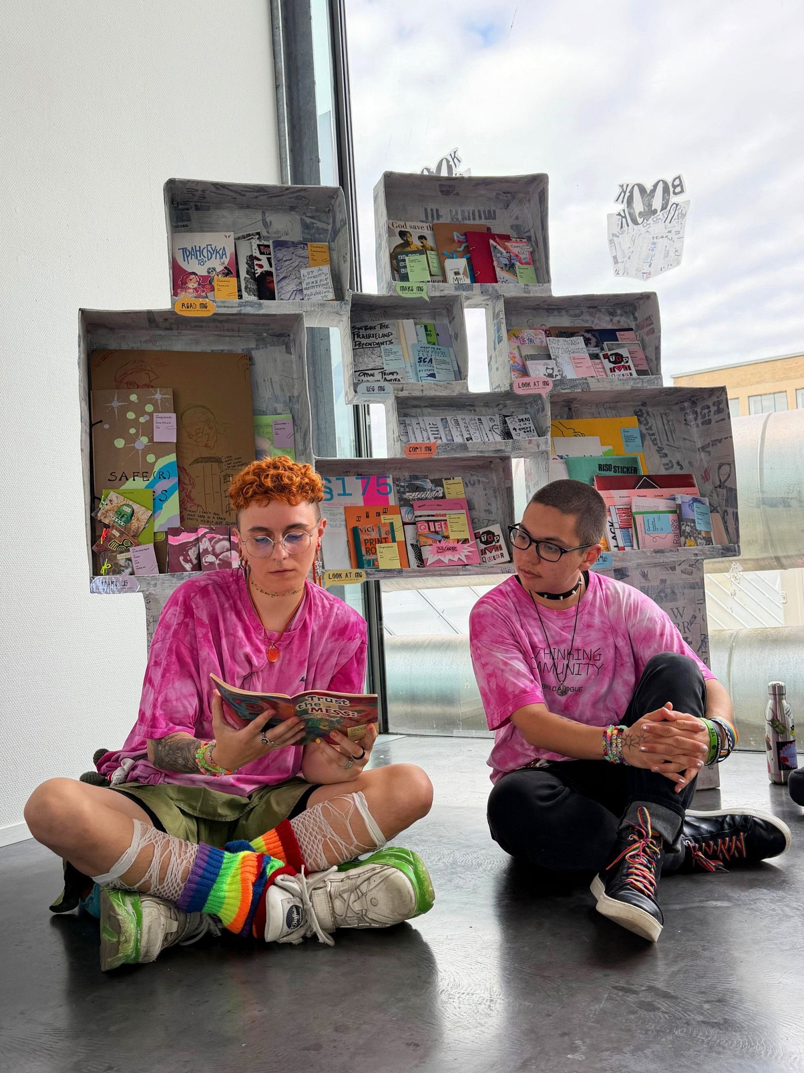 two people sitting infront of a zine shelf and reading a zine